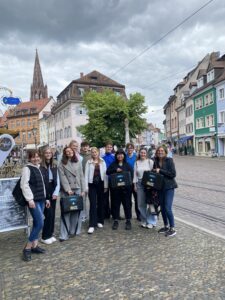 Eine Gruppe junger Menschen steht gemeinsam auf einem gepflasterten Platz in einer historischen Altstadt. Im Hintergrund sind bunte Häuser, ein Kirchturm und ein bewölkter Himmel zu sehen.