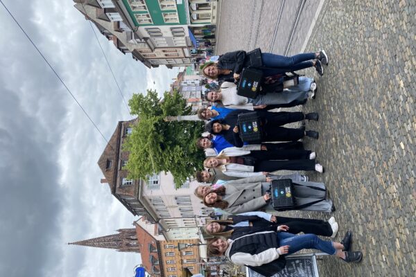 Eine Gruppe junger Menschen steht gemeinsam auf einem gepflasterten Platz in einer historischen Altstadt. Im Hintergrund sind bunte Häuser, ein Kirchturm und ein bewölkter Himmel zu sehen.